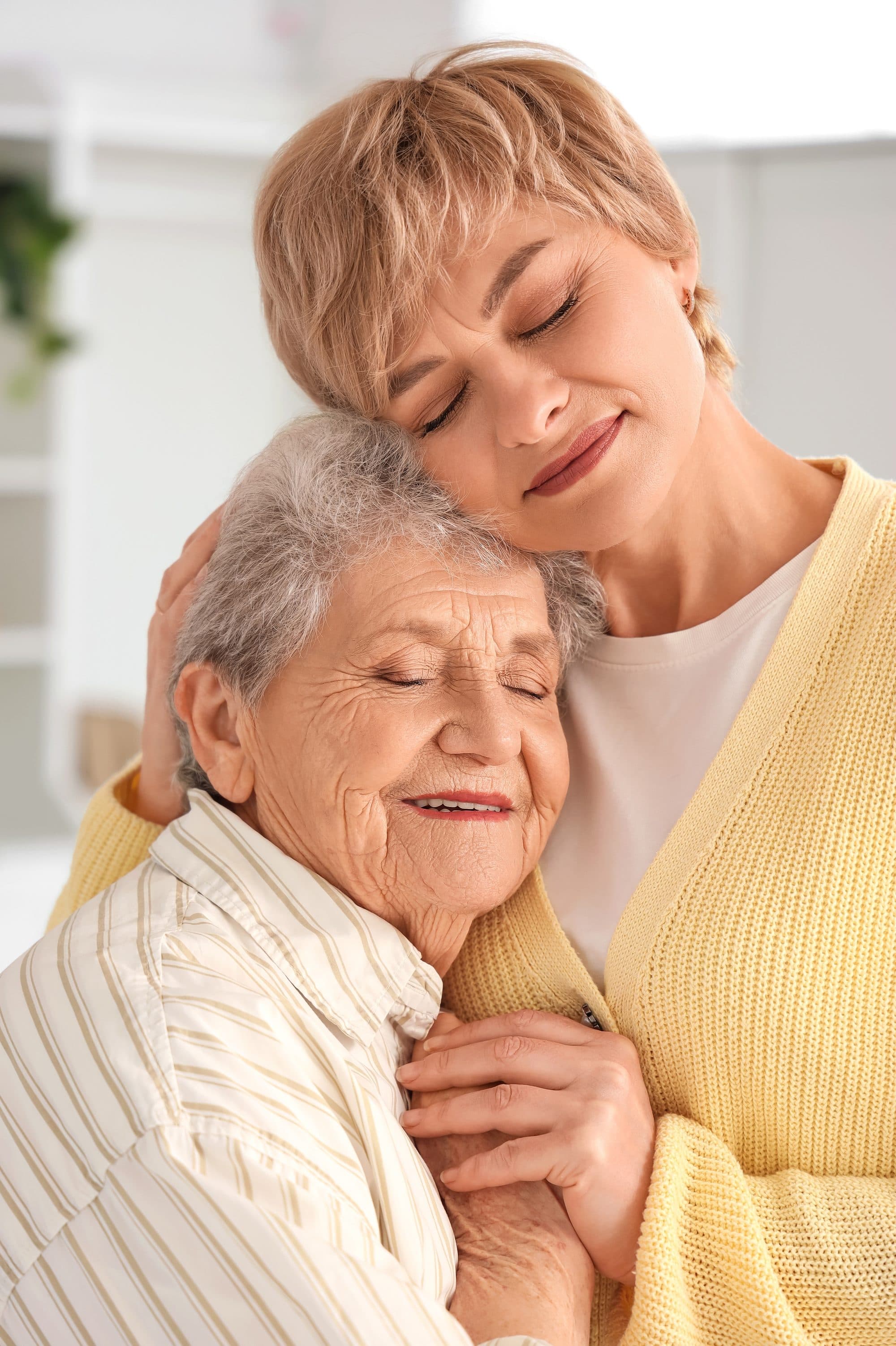 An adult daughter hugging her smiling elderly mother at home.