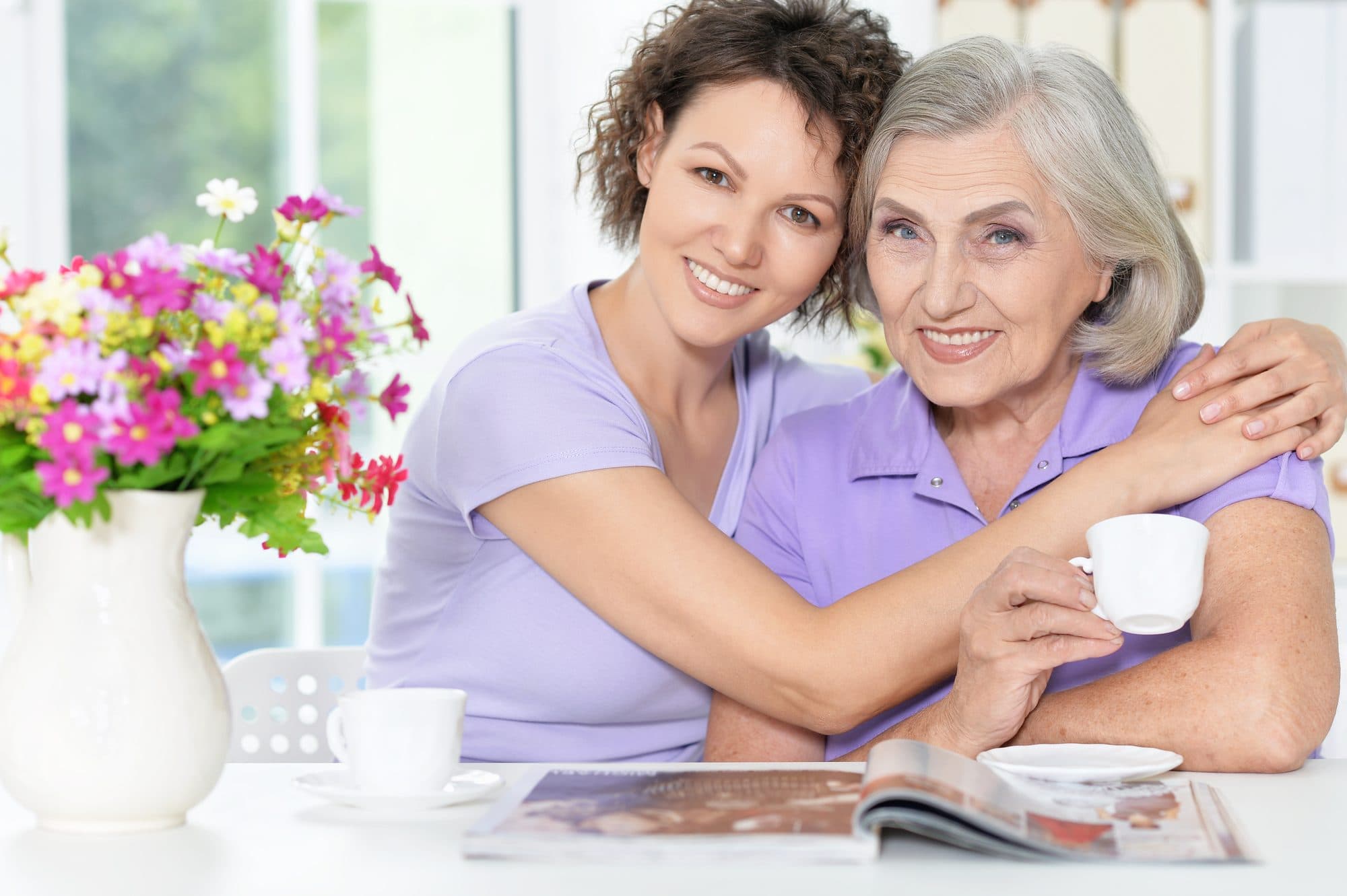 A mother and her adult daughter sharing tea at the kitchen table, smiling.