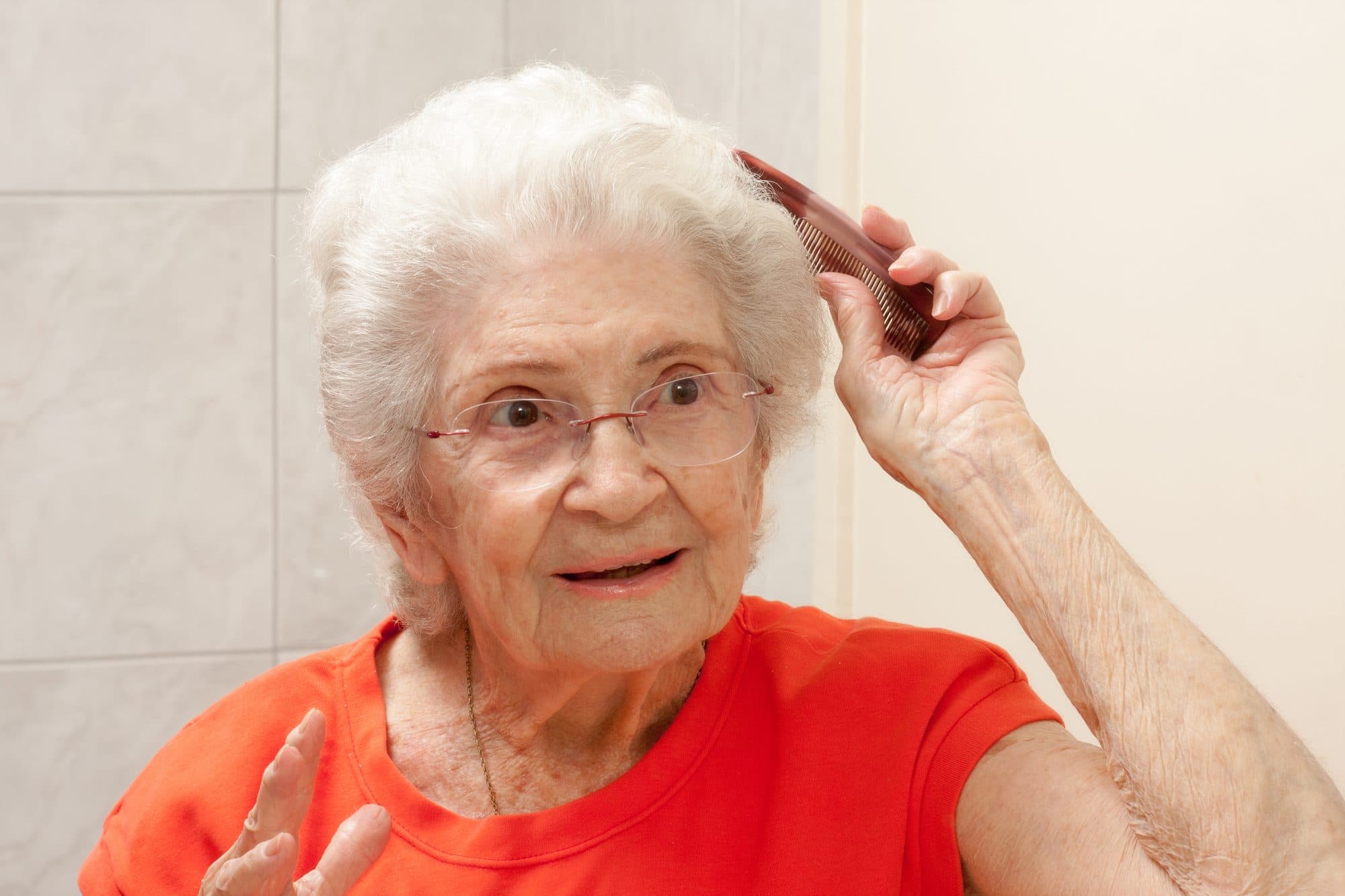 An elderly woman in a bright bathroom, brushing her hair and smiling.